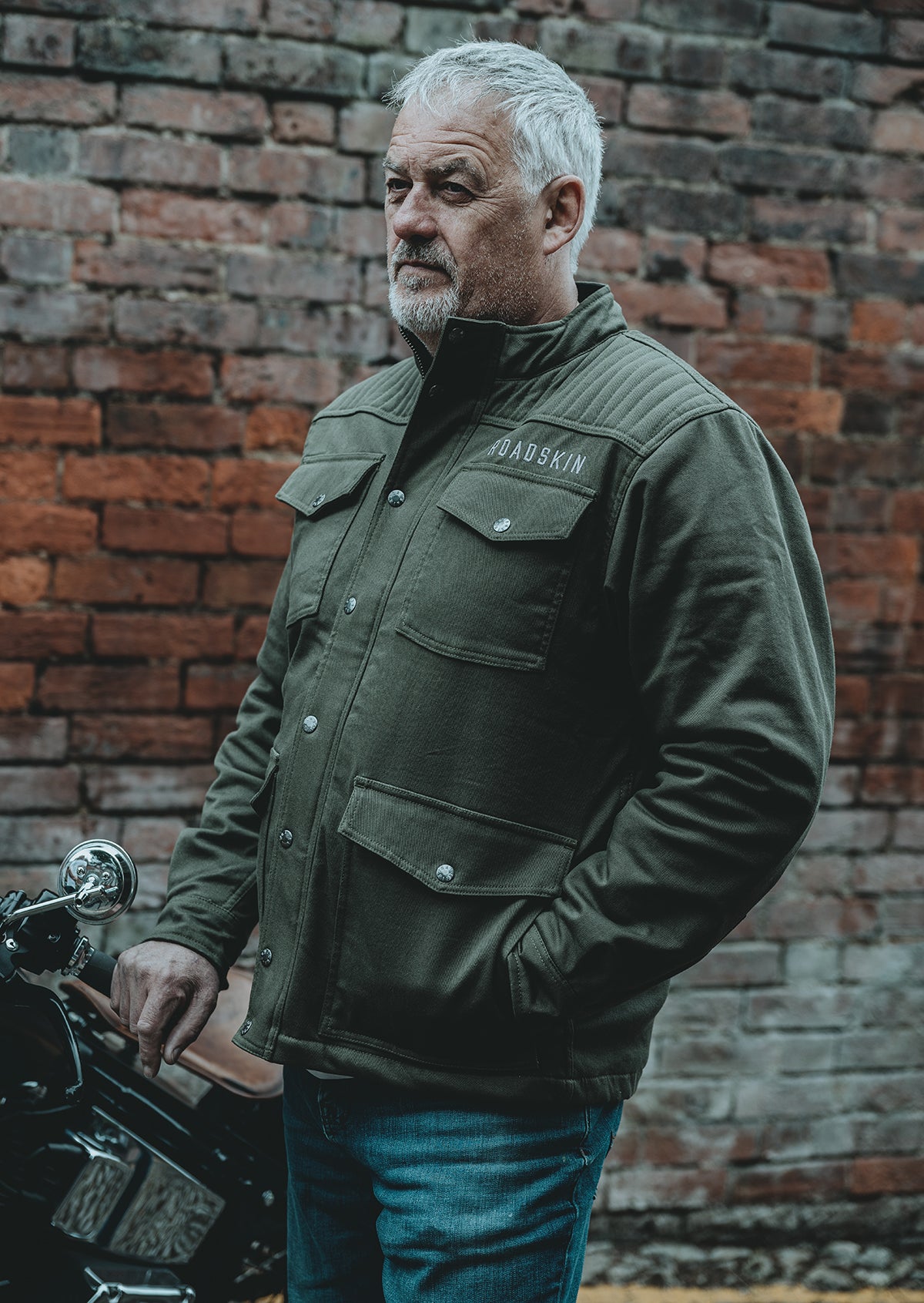 Man wearing a green motorcycle jacket standing next to a motorcycle against a brick wall.