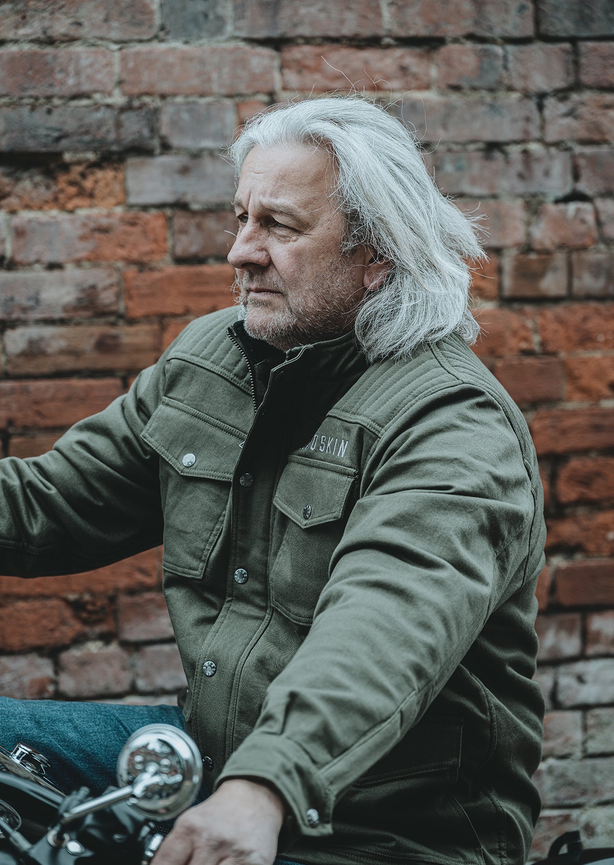 Man with white hair wearing a green motorcycle jacket sitting on a motorcycle against a brick wall.