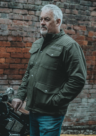Man wearing a green motorcycle jacket standing next to a motorcycle against a brick wall.